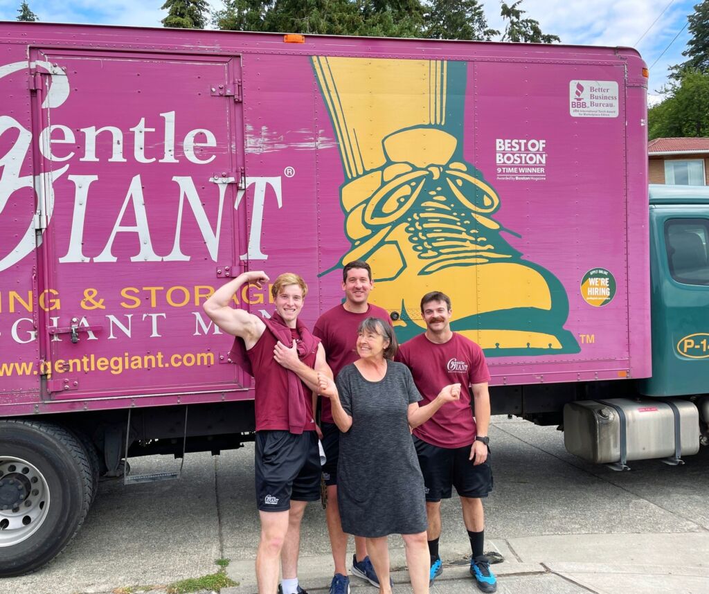 Three Gentle Giant movers dressed in maroon tshirts and black shorts are standing with a female customer in front of the purple Gentle Giant moving truck.