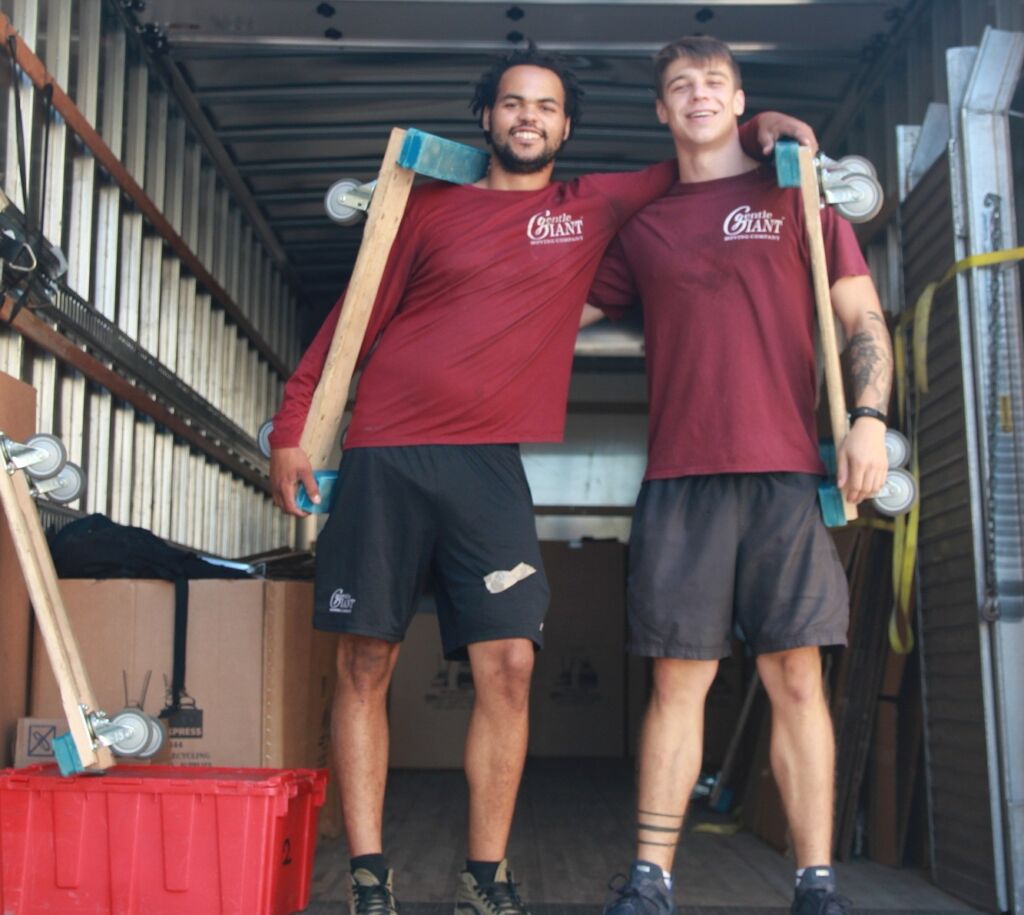 Two Gentle Giant movers are standing together, an arm is placed on top of each other's shoulder. They are standing and posing for a picture at the back of a moving truck that is open.