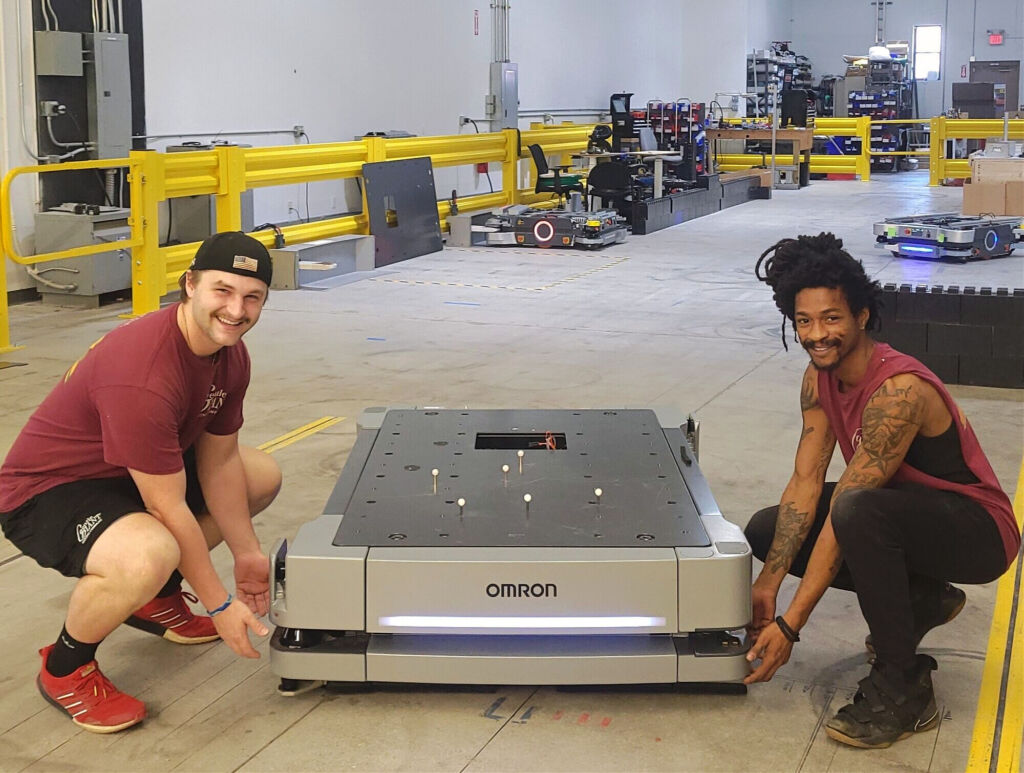Two Gentle Giant Moving Company team members crouch down and smile while handling an Omron autonomous mobile robot in a warehouse setting. Both are dressed in maroon Gentle Giant-branded athletic wear. The warehouse has yellow safety railings, various robotic equipment, and workstations in the background.