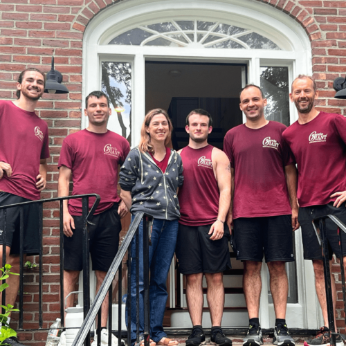 Five Gentle Giant movers are standing with a female customer at her front door entrance. Her home is a brownstone and she has a white front door. The movers are dressed in maroon t-shirts and black shorts.